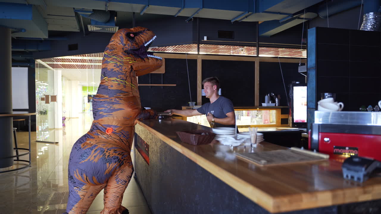 Person in dinosaur inflatable costume visiting the café. Orange dinosaur comes up to a bar stand and is given a menu by the bartender.