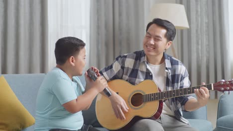 asian father playing the guitar for his son singing into microphone on sofa at home