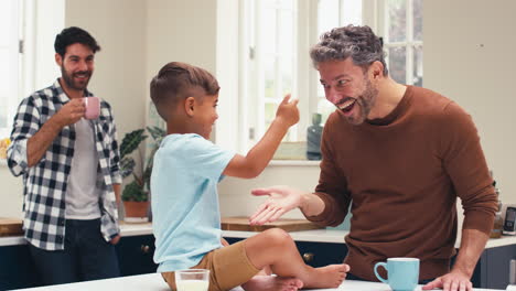 Familia-Del-Mismo-Sexo-Con-Dos-Padres-En-La-Cocina-Con-Un-Hijo-Sentado-En-El-Mostrador-Chocando-Los-Cinco-Con-Los-Padres