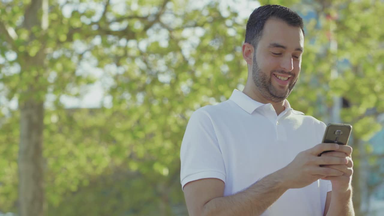 un joven sonriente paseando por el parque con un teléfono inteligente en las manos.