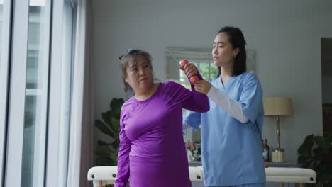 Asian-female-physiotherapist-helping-female-patient-exercise-arms-with-dumbbells-at-surgery