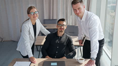 man in glasses sitting at desk and his colleagues standing on both sides. man closes laptop and all three look at camera smiling.