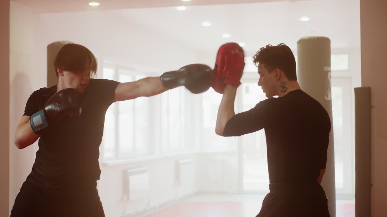 Boxer in black gloves faces partner holding red pads during training session inside gym, demonstrating combat stance, readiness, focus, energy, discipline, power, precision, coordination
