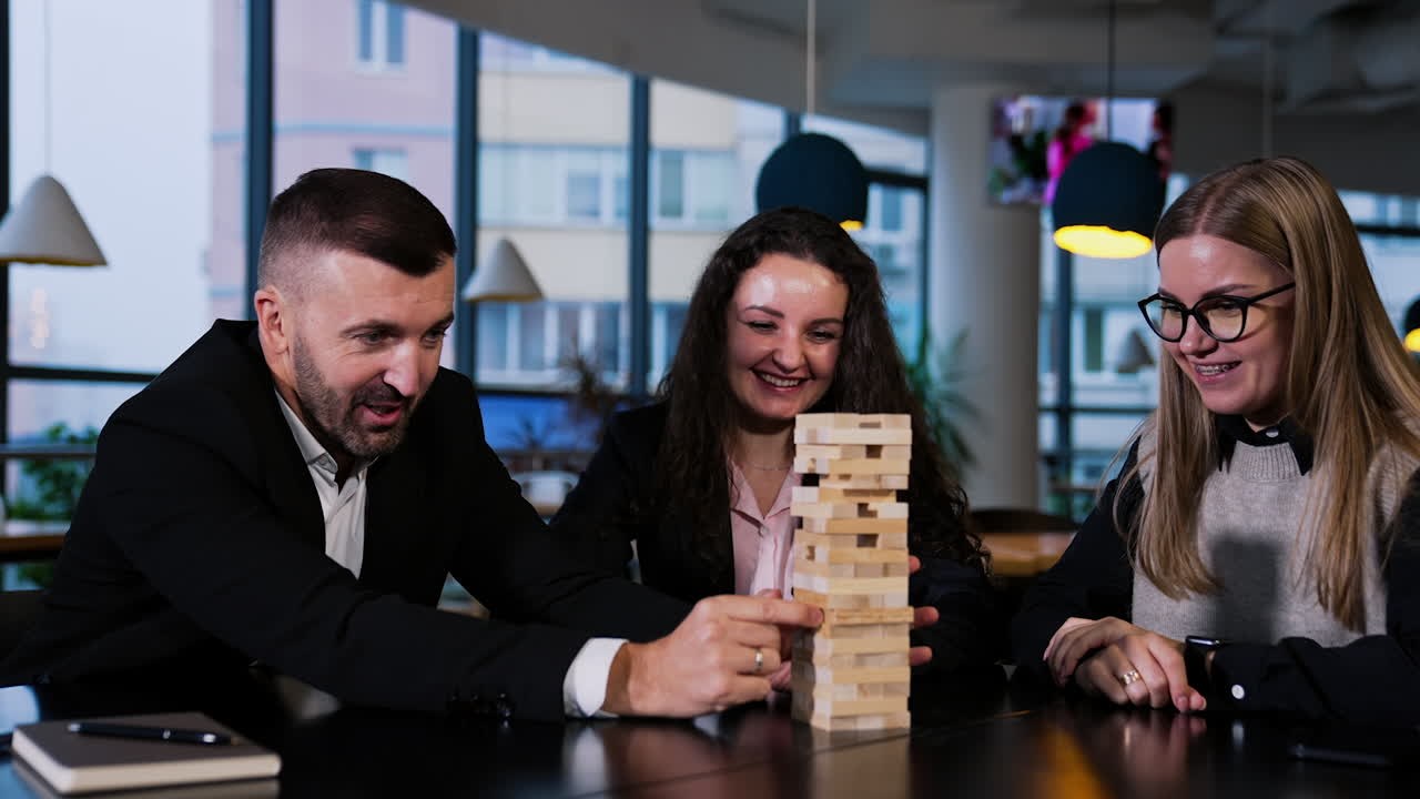 Smiling cheerful people play jenga sitting at the table. Bearded man pulling block while women laugh and comment.
