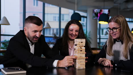 smiling cheerful people play jenga sitting at the table. bearded man pulling block while women laugh and comment.