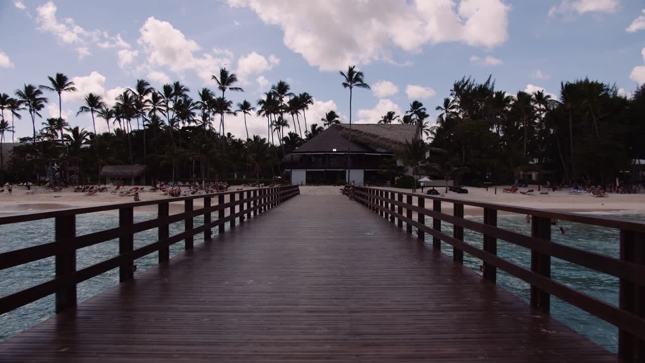 Dolly in shot on wooden pier toward tropical beach; palms against skyline