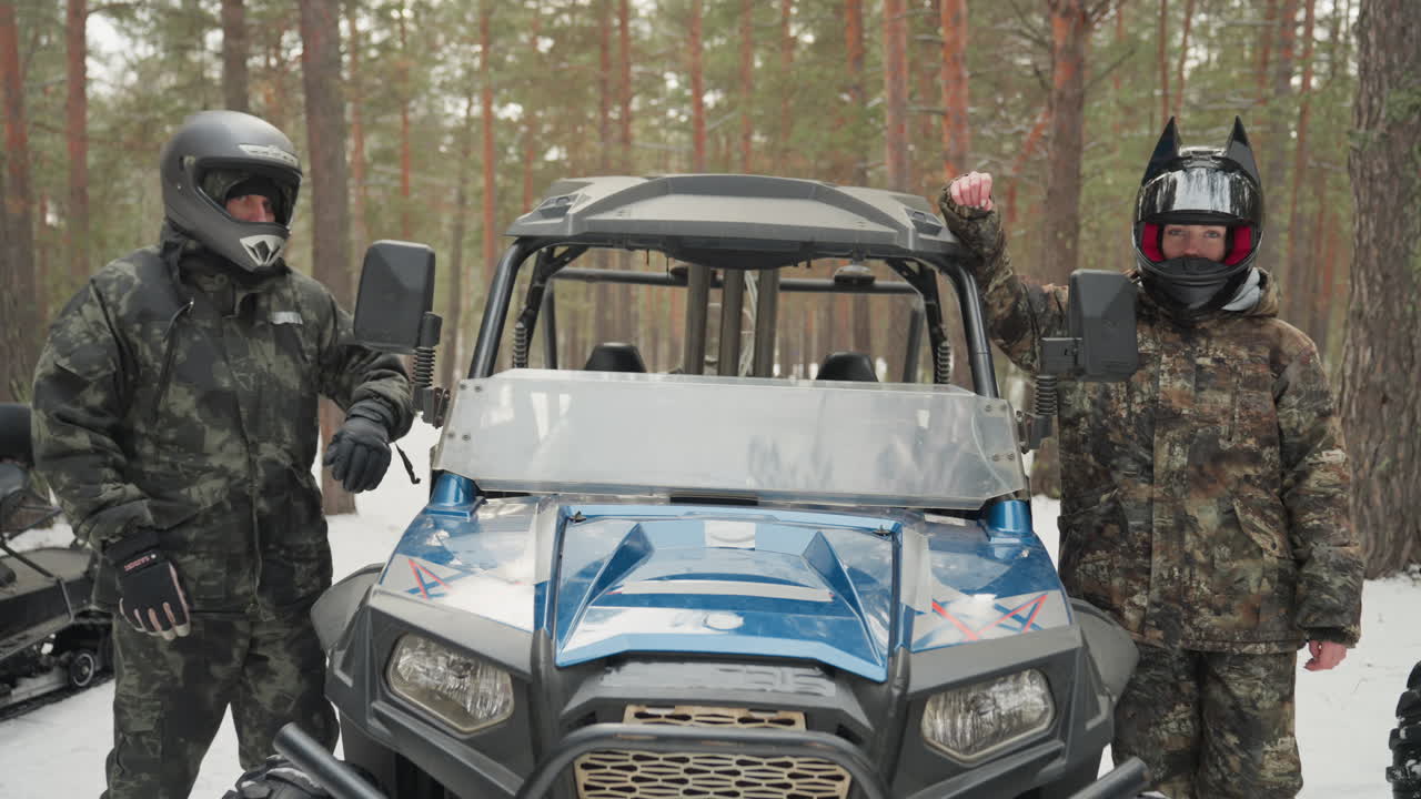 Nærbillede af blå UTV med ryttere, der forbereder sig, to hjelmklædte personer i camouflage- og sort udstyr står ved siden af maskinen, snescooter og sort ATV i baggrunden, vinterfyrreskov, frossen jord, klar position.