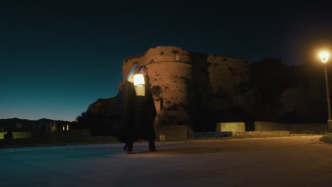 woman with cloak and lantern spins in a circle pirouette at night near a castle