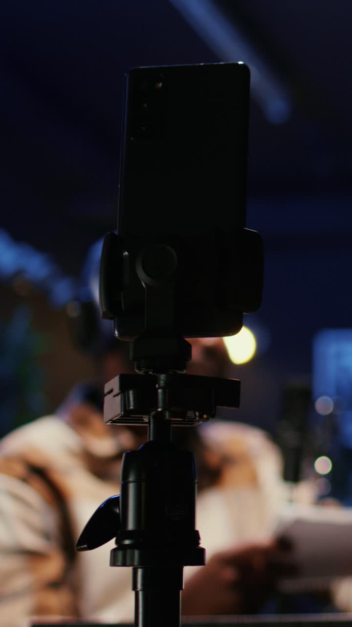 Vertikal video. Smartphone på stativ optager vloggershows. Værten i sløret baggrund interviewer gæst.