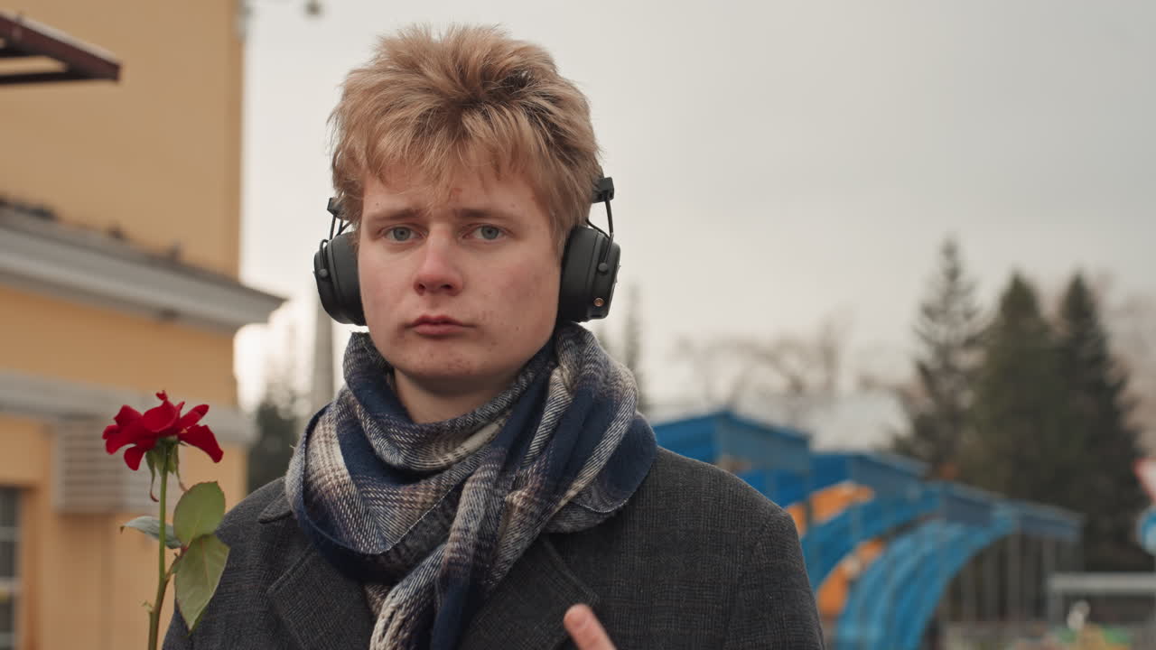 White man headphones smiling with rose, small urban station backdrop, scarf and coat, playful creator energy as he offers flower and flashes cheeky finger, warm expression, candid busstop portrait