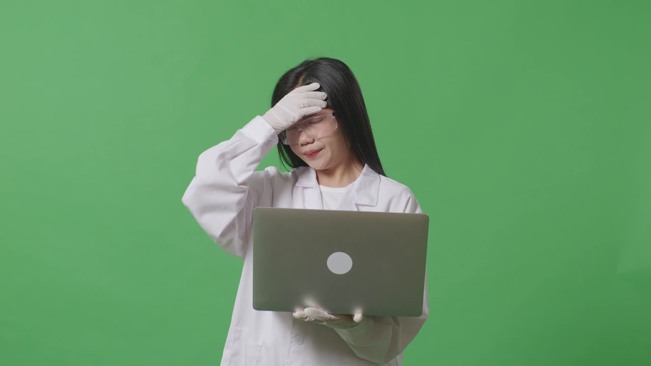 científica asiática con dolor de cabeza mientras está de pie y trabajando en una computadora portátil en el laboratorio de fondo de pantalla verde