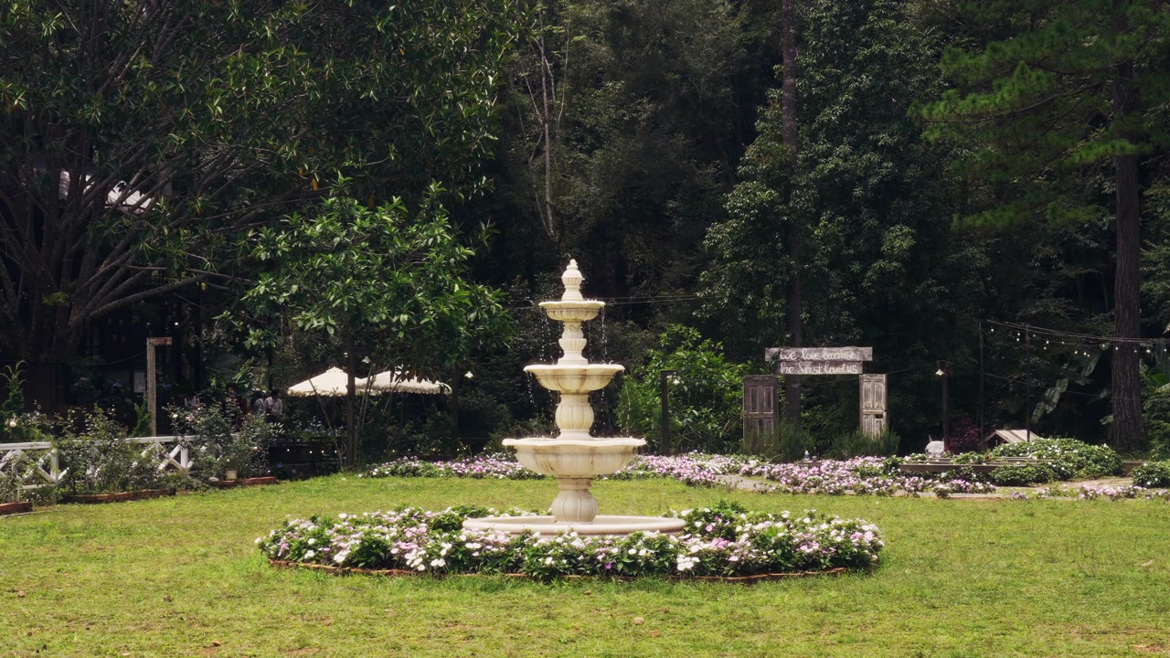 緑豊かな庭園で水しぶきを上げる古典的な段状の石の噴水。色とりどりの花壇と鬱蒼とした森が背景にあり、平和で穏やかな雰囲気を作り出している。