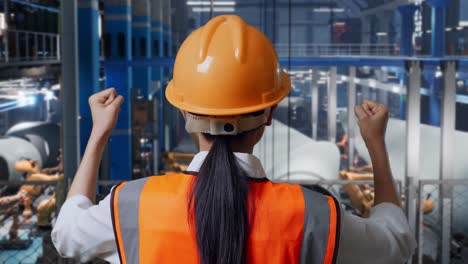 vista cercana de atrás de una ingeniera asiática con casco de seguridad de pie levantando las manos celebrando trabajando en una fábrica de fabricación de turbinas eólicas