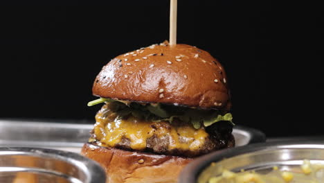 Reverse-Dolly-Zoom-on-Savory-Cheeseburger-with-Sesame-Seed-Bun-and-Sous-Plates-in-Front,-Black-Background,-Landscape-View