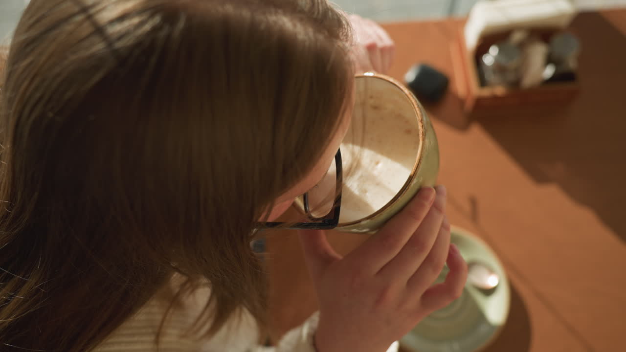 Top down view of woman wearing cozy sweater lifting ceramic coffee cup from table, soft shadows cast by morning sunlight, saucer and spoon remain on wooden surface