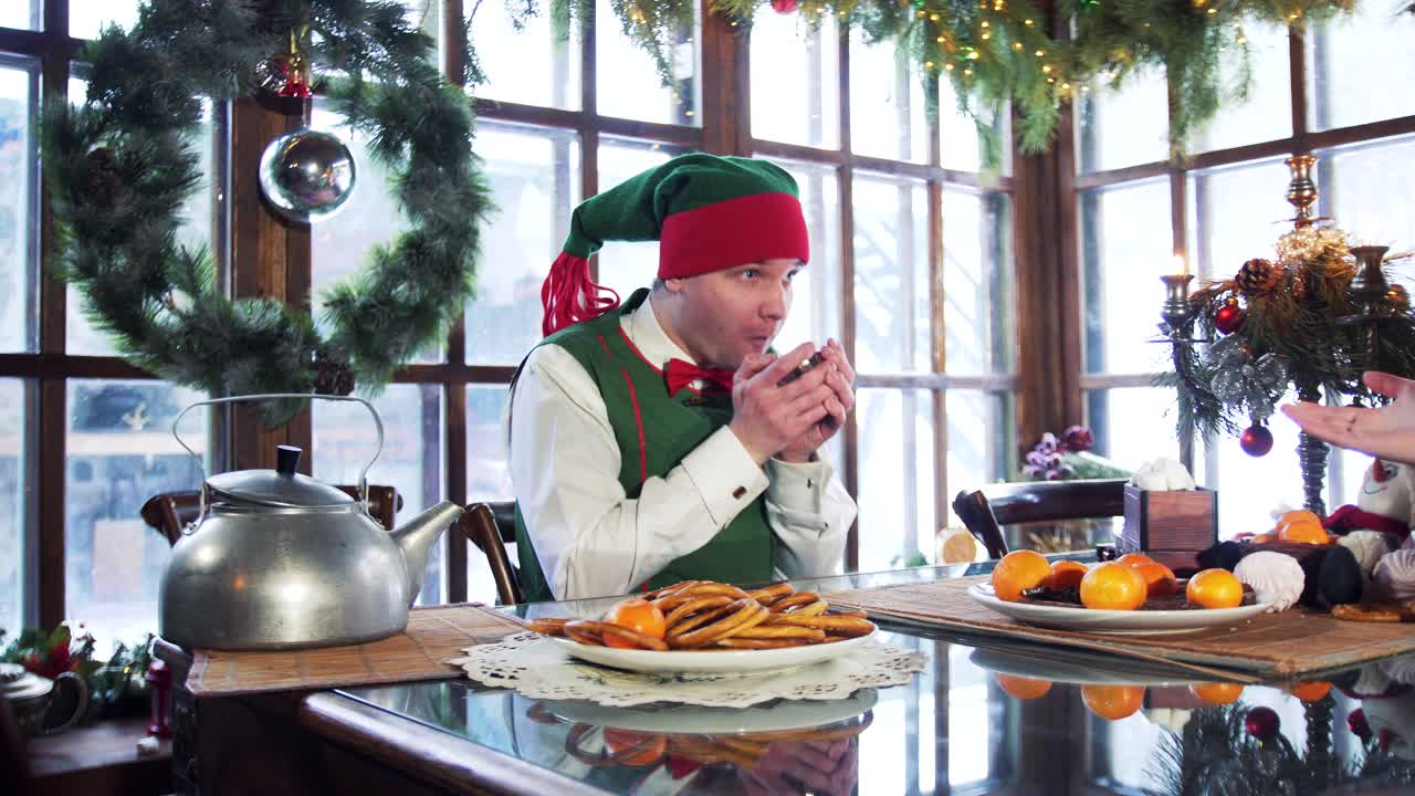 Elf male with cup sitting at table on Christmas background. Funny elf in green costume gets up from a table