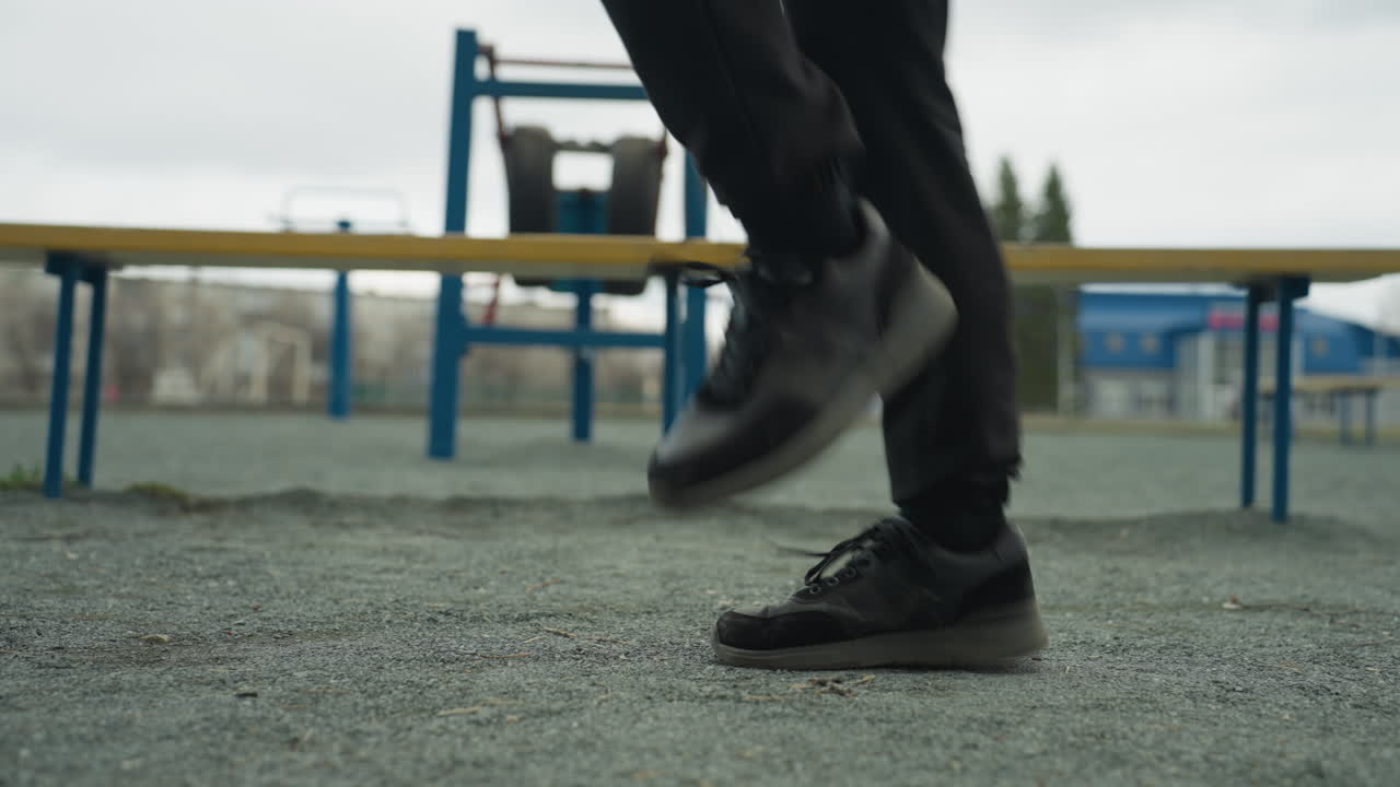Close-up of a person's legs jogging rapidly, wearing black jeans and black sneakers, with benches around and distant houses visible in the background