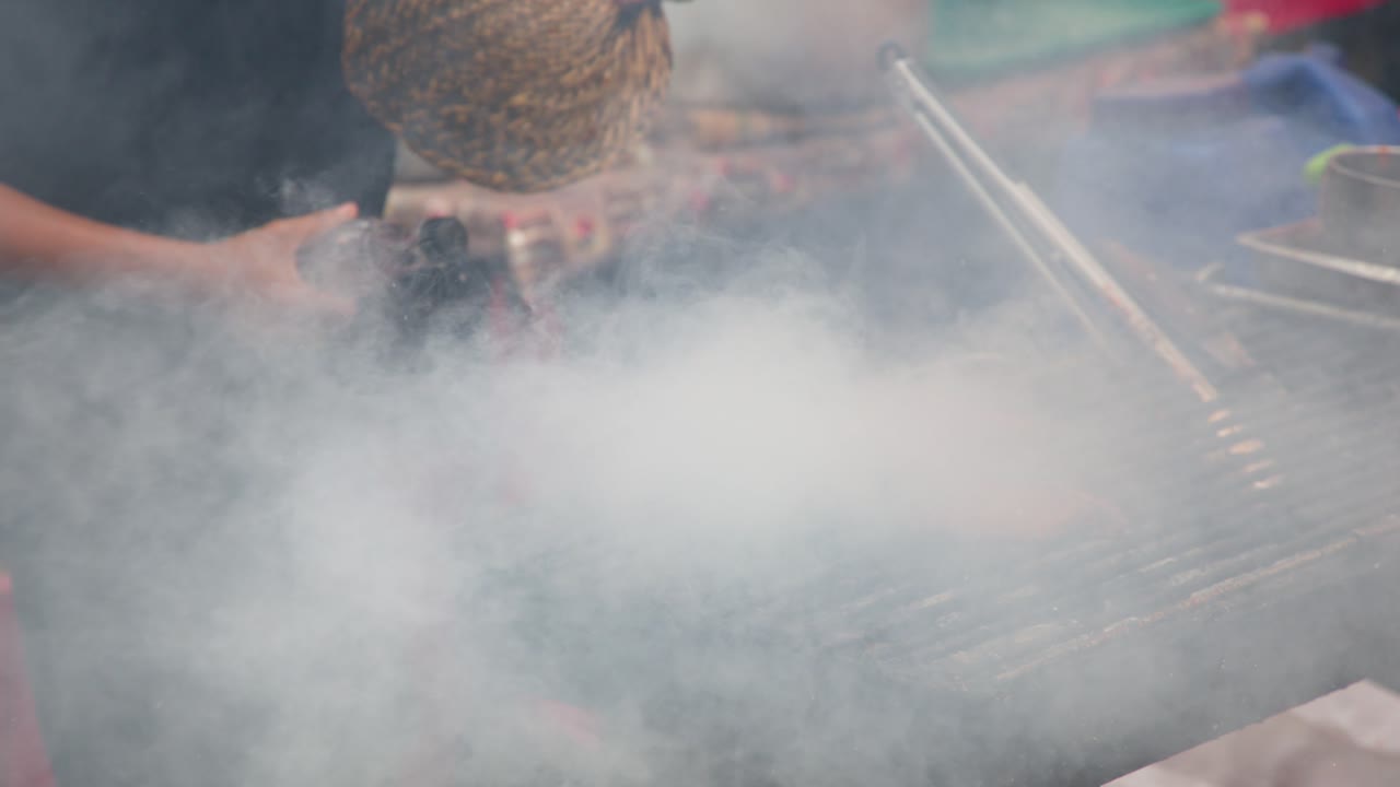 Un venditore ambulante griglia spiedini di pollo sulla brace, creando denso fumo bianco e fiamme.