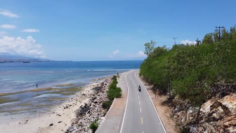 aerial drone of a beautiful coastal road and the ocean with a scooter motorbike driving around a blue sky sunny day on tropical island