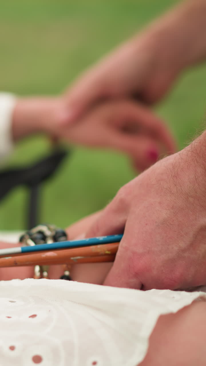 In a peaceful outdoor scene, an artist in a checkered shirt gently holds a paintbrush while carefully adjusting the hand of a seated woman wearing a white dress. The woman's hand, adorned with beads