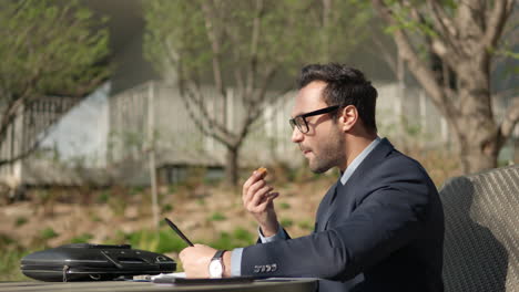 Young-Adult-Man-in-Suit-Snacking-Outdoors-in-a-Park-While-Reviewing-Paper-Notes,-Highlighting-Important-Text-Sitting-in-a-Chair-by-the-Table