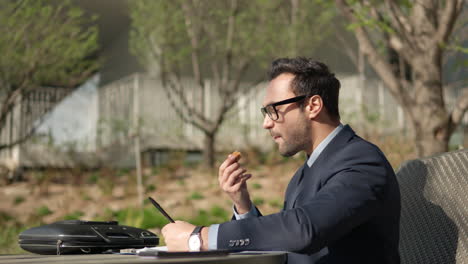 young adult man in suit snacking outdoors in a park while reviewing paper notes, highlighting important text sitting in a chair by the table