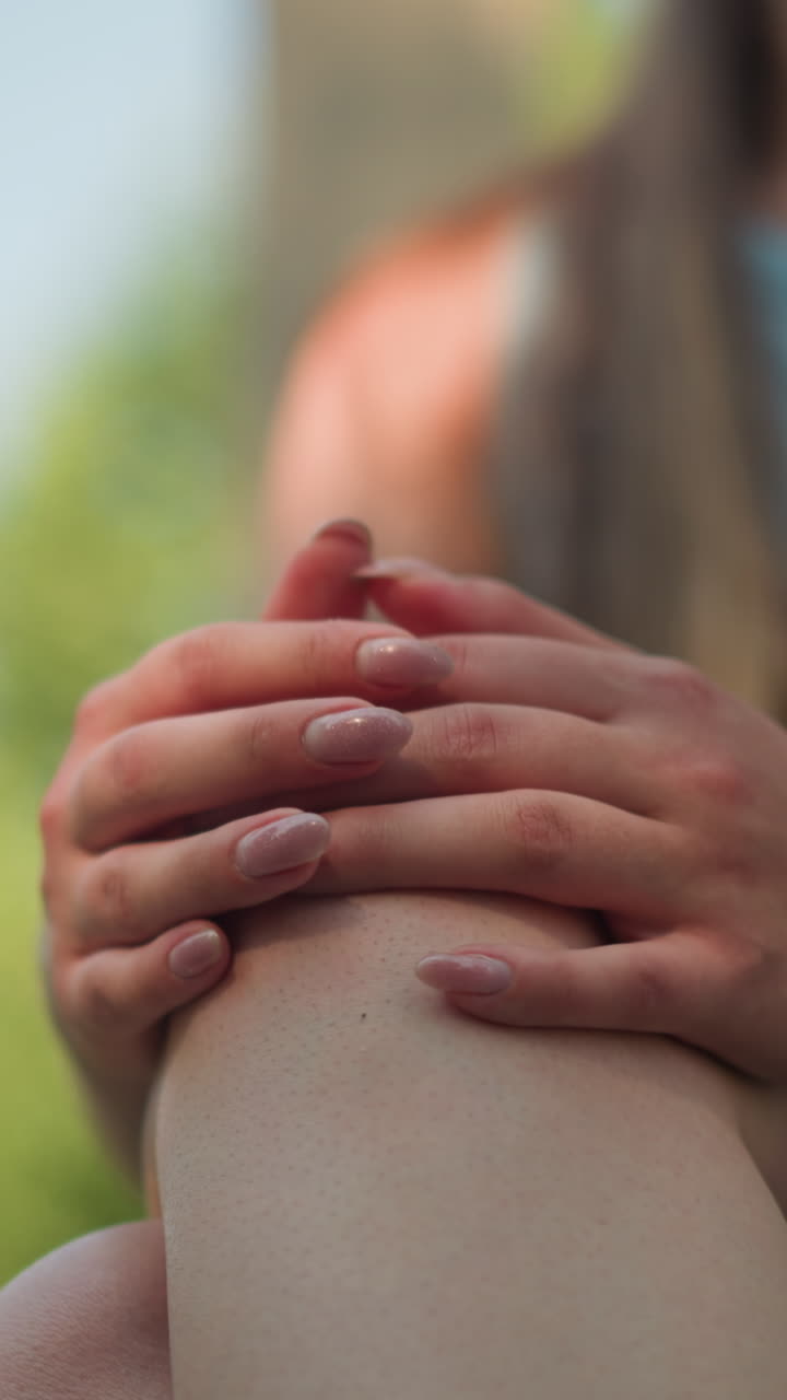 vista parcial de una mujer joven sentada al aire libre con las piernas cruzadas, las manos juntas en la rodilla, haciendo movimientos suaves con los dedos, usando un brazalete de mano negro, usando una camiseta cian, bajo la suave luz del sol