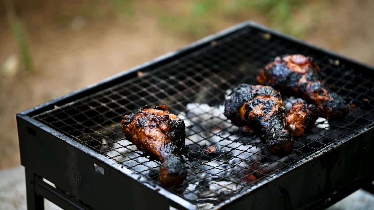 Slow-motion cinematic shot shifting from blur to focus on burnt chicken drumsticks cooking over low flames on a BBQ grill. Highlights rich food textures, smoky outdoor cooking, and authentic grilling