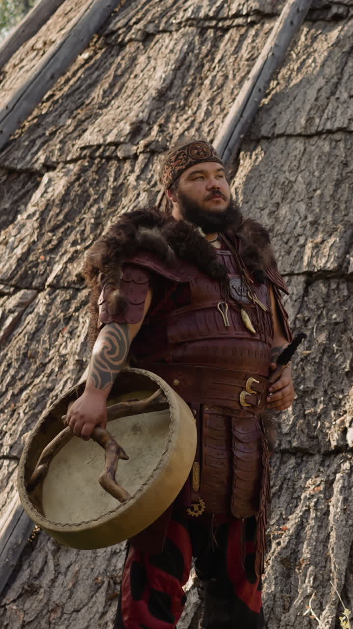 Yakut shaman in traditional costume holds tambourine standing by conical ail building. Actor plays Altai wizard. Ethnic festival atmosphere