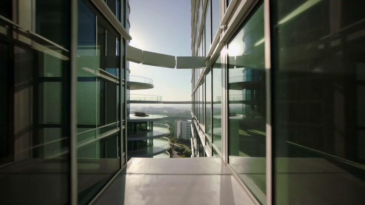 A câmera começa a fazer um movimento panorâmico para baixo em uma passarela estreita em direção a uma passarela elevada entre torres, revelando o horizonte.