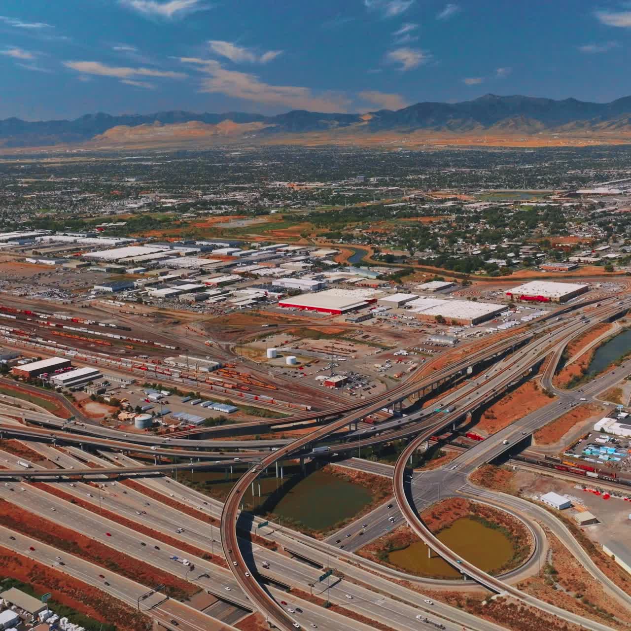Industrial zone in Salt Lake City with lots of intertwining highways. Residential part of the city with thick greenery at backdrop. Utah mountains at backdrop