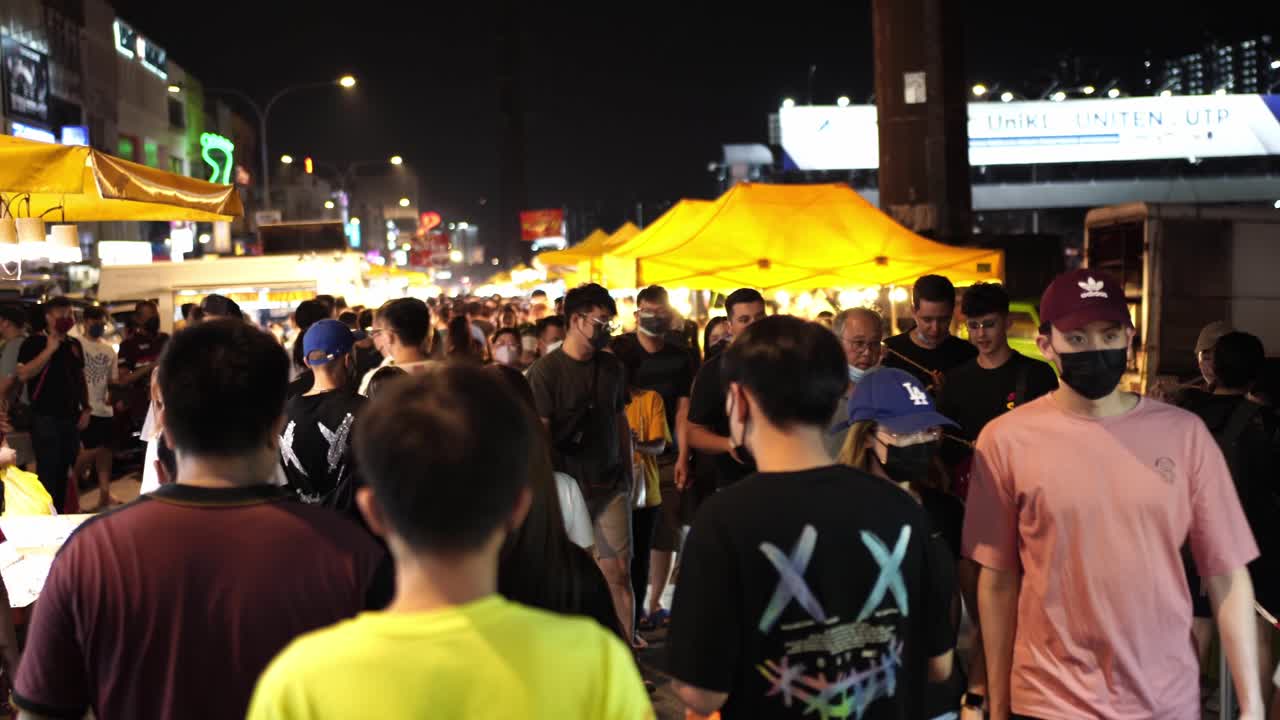 People walking through a busy night market in Kuala Lumpur, Malaysia, wearing face masks, enjoying street food, with bright street signs and yellow marquees illuminating the scene.