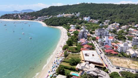 Luftdrohnenszene-Des-Touristischen-Strandes-In-Florianopolis-Mit-Vielen-Sommerhäusern-Hotels-Mit-Blick-Auf-Das-Meer-Städtischer-Strand-Mischt-Sich-Mit-Vielen-Menschen,-Die-Die-Sonne,-Den-Sand-Und-Das-Meer-Genießen-Santa-Catarina