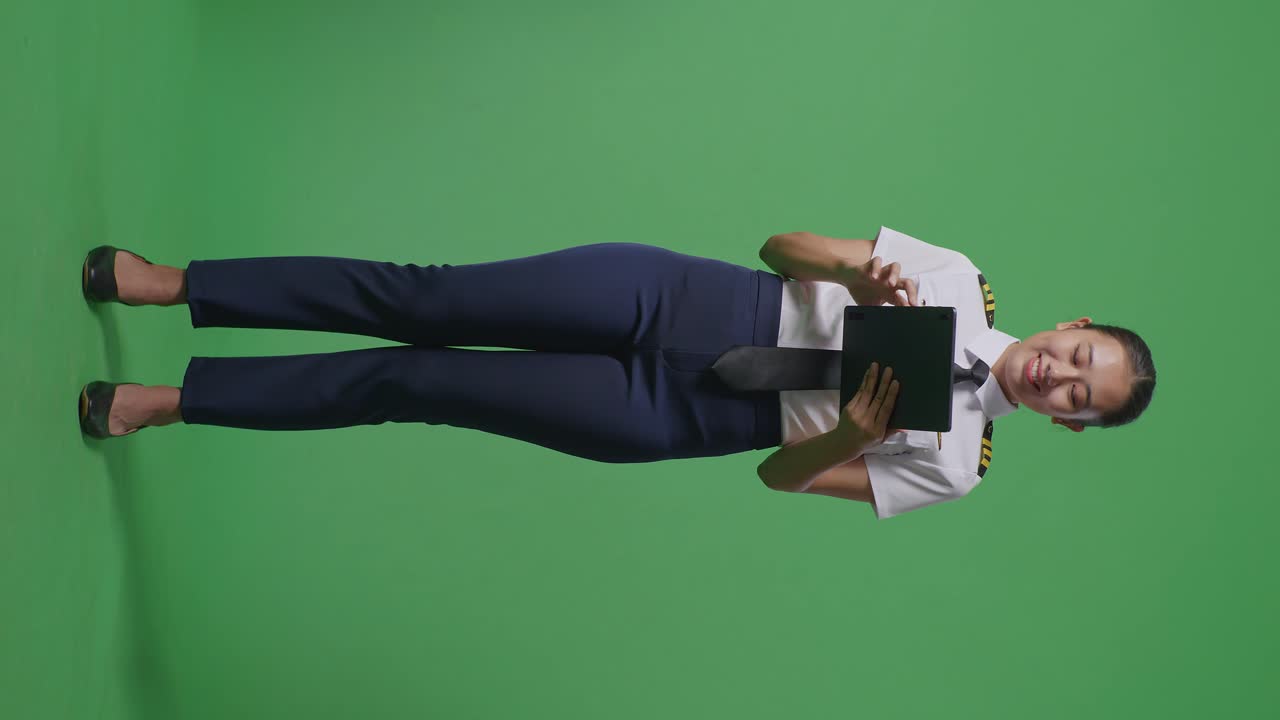 cuerpo lleno de piloto mujer asiática usando una tableta y apuntando a un lado mientras está de pie en el estudio de fondo de la pantalla verde