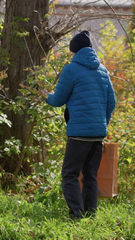 niño joven con una chaqueta azul y gorra negra caminando hasta un árbol, arrancando hojas, y poniéndolas en una bolsa de papel, con un edificio en el fondo y la vegetación que lo rodea