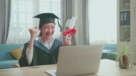Asian Woman Celebrating With University Certificate To The Family During An Online Video Call. Pretty Female Graduate Wearing A Graduation Gown And Cap Sitting On The Living Room
