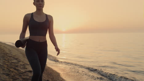 athletic woman walking beach holding sport mat at sunset. girl going exercising.