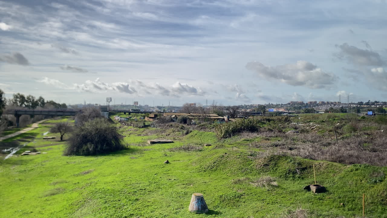 Prati verdi lussureggianti e un sentiero allagato in una pianura fluviale urbana, con edifici cittadini in lontananza sotto un cielo nuvoloso.