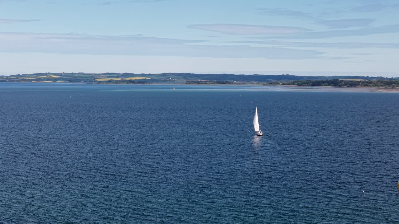 Aerial drone view of a white sailboat gliding across calm blue waters with coastline in the distance