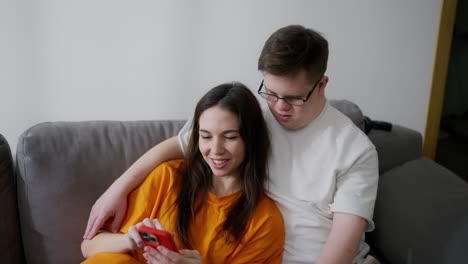 Brother-with-Downs-Syndrome-sitting-on-sofa-with-his-sister,-she-is-using-mobile-phone