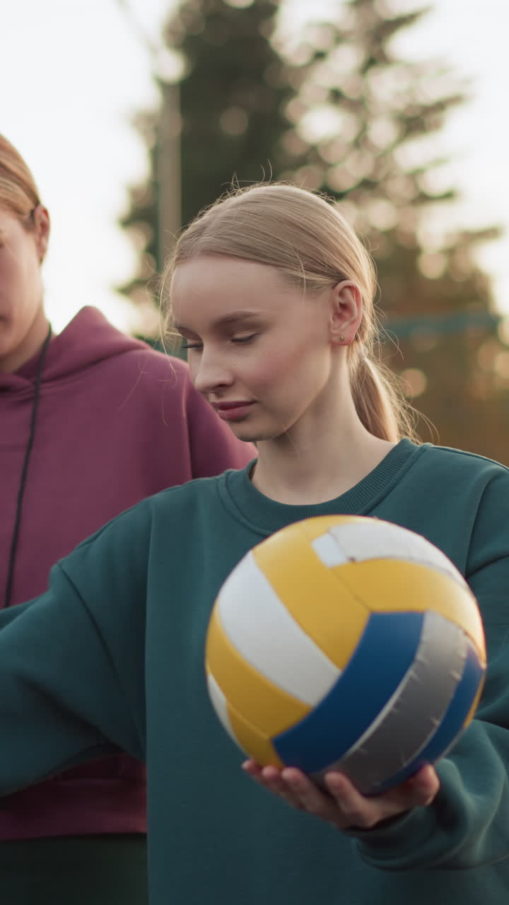 배구 강사가 초보자에게 서브 기법을 보여 주고, 그 다음에는 공을 서브하도록 허용합니다. 둘 다 손에 배구 공을 들고 야외 코트에 서 있습니다.