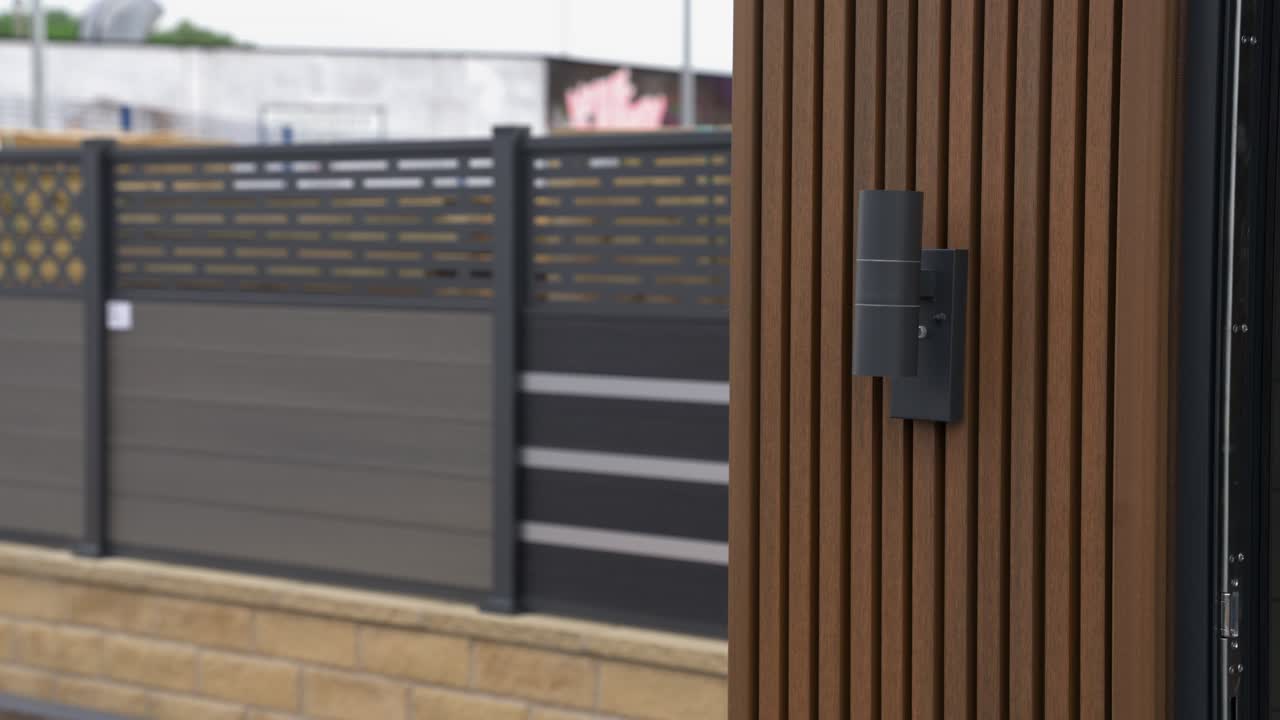an up and down light sitting on the end of brown luxury cladded garden room in a garden showroom with different examples of fencing in the background