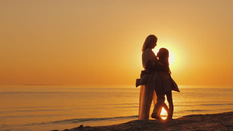 Mom-Gently-Hugs-The-Child-Against-The-Background-Of-The-Sea-And-The-Setting-Sun-Silhouettes-Of-Woman