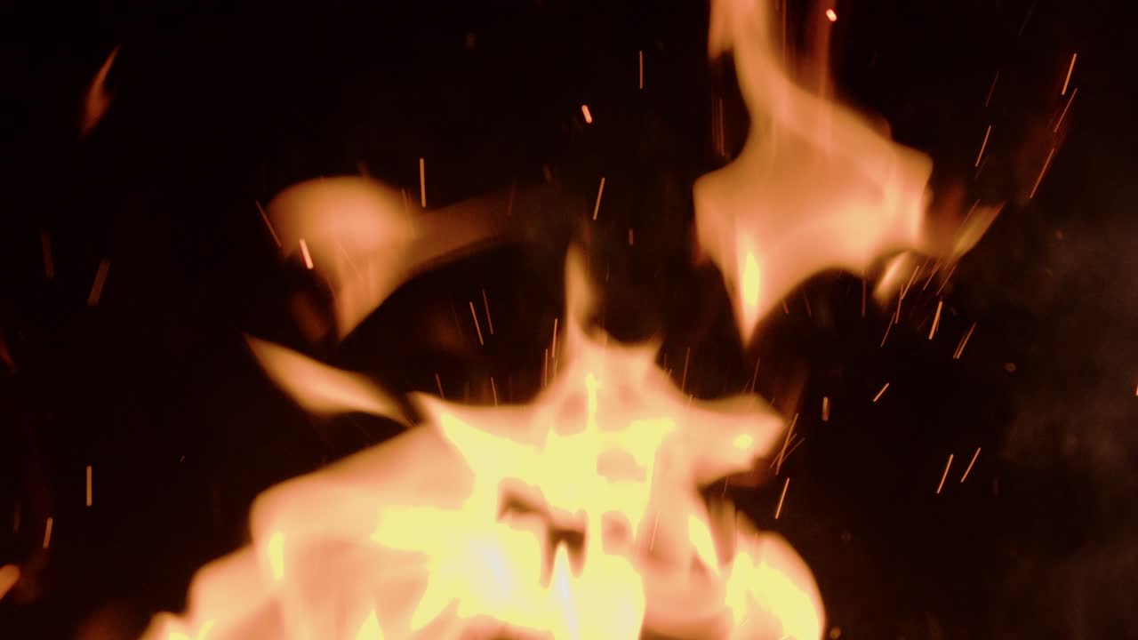 Dynamic close-up shot of slow motion fire flames and glowing sparks rising against a dark background. Ideal for use in cinematic scenes, survival themes, or dramatic visual effects