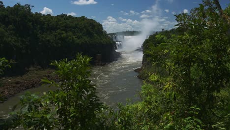 slow motion river flowing from huge aggressive rainforest waterfall, slo mo water stream heading through tall thick jungle scenic location, bright sunny weather conditions in iguacu falls