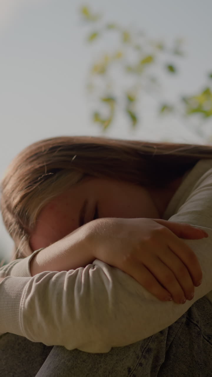 Thoughtful young woman sits on grassy ground with folded arms, resting her head on her hand as sunlight softly illuminates her face, surrounded by greenery and blurred background