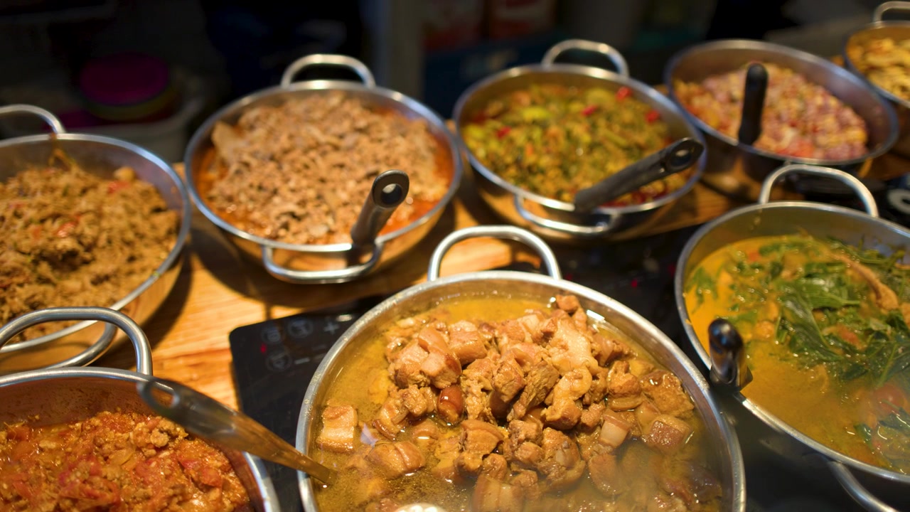 Inquadratura statica dall'alto di vari stufati e curry di maiale thailandesi piccanti in pentole di metallo.