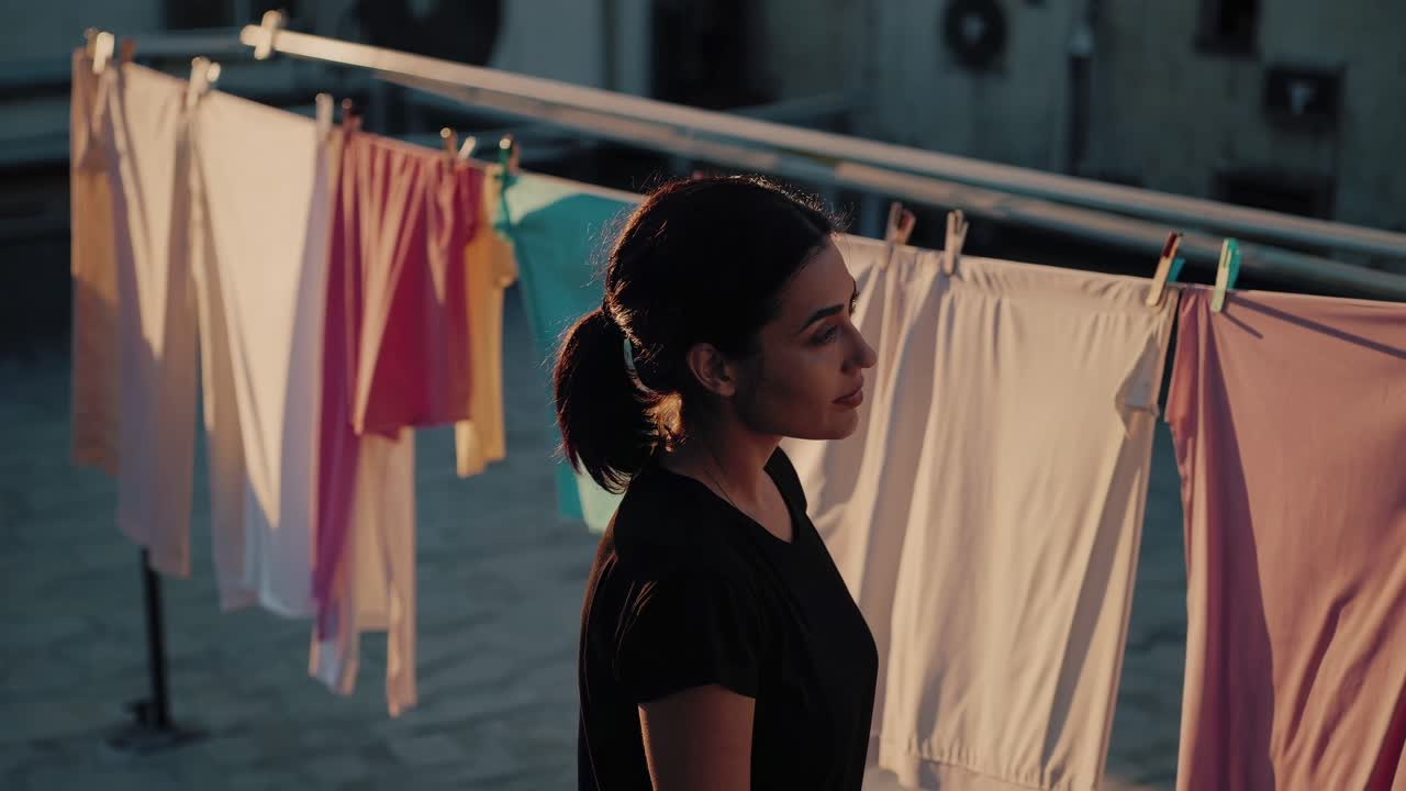 Young woman standing on rooftop at sunset, hanging freshly washed clothes, experiencing moment of calm amid urban landscape, embodying simple domestic tranquility