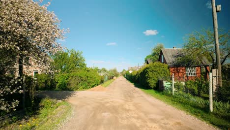 Aufnahmen-Einer-Sandigen-Schotterstraße,-Gesäumt-Von-Einem-Blühenden-Baum,-Einem-Garten-Und-Einem-Sommerhaus-Unter-Einem-Blauen-Himmel-Mit-Sonnenlicht-Und-Wolken