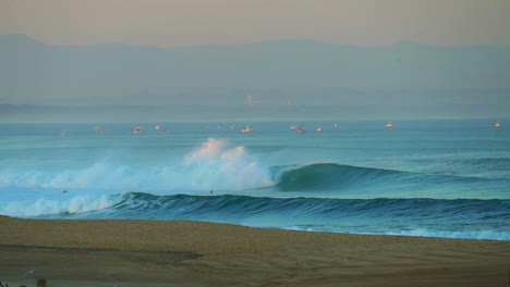 Cinematic-pan-stunning-early-morning-huge-waves-glassy-swell-surf-close-up-with-sailboats-Hossegor-Seignosse-France-pink-purple-orange-sunrise-sunset-on-beach-mountain-coast-Sailboats-at-bay